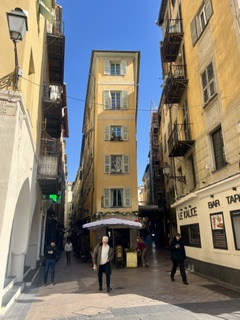 A sunlit narrow street lined with tall ochre buildings and shuttered windows.