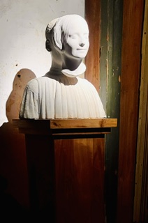 A marble bust of a young woman on a wooden pedestal in warm gallery light.