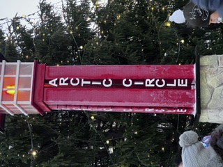 A red and white Arctic Circle marker glowing among snow-dusted evergreens and string lights.